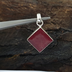 Puede incluir: Colgante de gema roja oscura facetada y cuadrada con engaste de plata. El colgante está sujeto con pinzas sobre un fondo de madera oscura y texturizada. La gema tiene forma geométrica y superficie pulida.