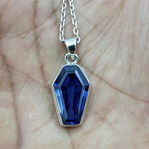 May include: A silver chain necklace with a pendant shaped like a coffin. The pendant features a large, faceted, dark blue gemstone.