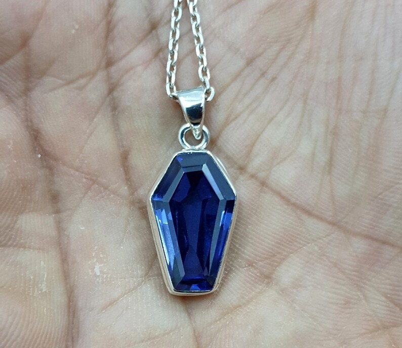 May include: A silver chain necklace with a pendant shaped like a coffin. The pendant features a large, faceted, dark blue gemstone.
