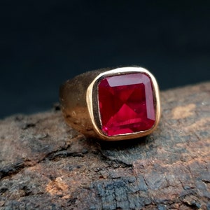 Pu&ograve; includere: Un anello in oro con una grande pietra preziosa rossa vibrante a taglio quadrato, incastonata in una fascia di metallo liscia e lucida. La montatura &egrave; spessa e solida, mettendo in risalto il colore cremisi profondo della pietra sfaccettata.