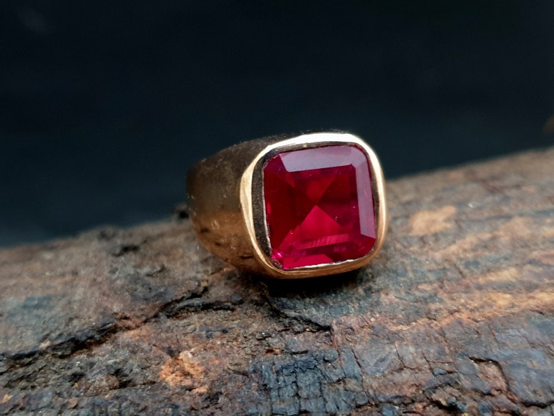 Pu&ograve; includere: Un anello in oro con una grande pietra preziosa rossa vibrante a taglio quadrato, incastonata in una fascia di metallo liscia e lucida. La montatura &egrave; spessa e solida, mettendo in risalto il colore cremisi profondo della pietra sfaccettata.