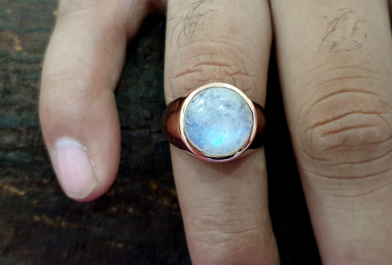 Puede incluir: Un anillo de plata con una gran piedra de luna redonda y blanca engastada en un engaste de bisel. El anillo est&aacute; en un dedo.