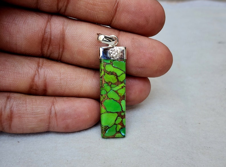Pu&ograve; includere: Un ciondolo rettangolare con una vibrante pietra verde e marrone, sormontato da un'ansa color argento. La pietra presenta un motivo naturale unico. Il ciondolo &egrave; tenuto in una mano.