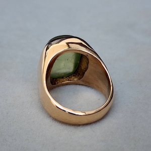 Puede incluir: Un anillo de oro con una gran piedra preciosa verde engastada en forma cuadrada. El anillo es de estilo sello con una banda ancha.