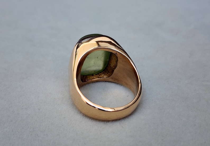 Puede incluir: Un anillo de oro con una gran piedra preciosa verde engastada en forma cuadrada. El anillo es de estilo sello con una banda ancha.