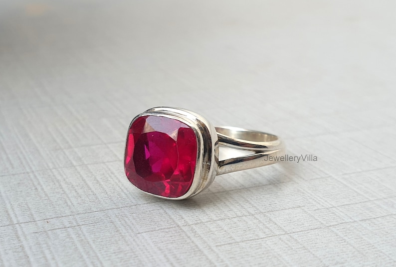 May include: A silver ring featuring a large, square-shaped, bright red gemstone. The ring has a double band with the text "JewelleryVilla" visible on the band.