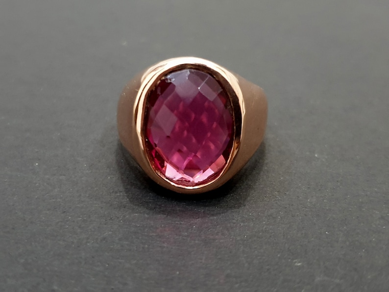 May include: A rose gold ring with a large, faceted, oval-shaped, pink gemstone.