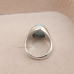 K&ouml;nnte beinhalten: Ein silberner Ring mit einem hellblauen ovalen Edelstein, der in einer Zargenfassung sitzt. Der Ring wird von hinten gezeigt.