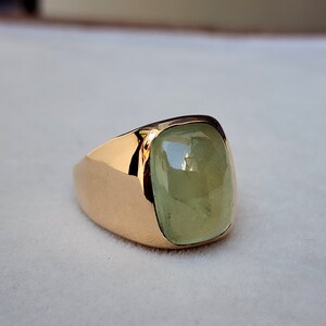 Puede incluir: Un anillo de oro con una gran piedra preciosa verde claro. El anillo tiene un engaste cuadrado y una banda ancha.