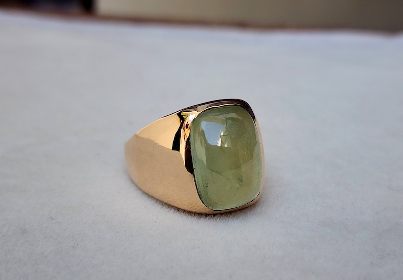 Puede incluir: Un anillo de oro con una gran piedra preciosa verde claro. El anillo tiene un engaste cuadrado y una banda ancha.