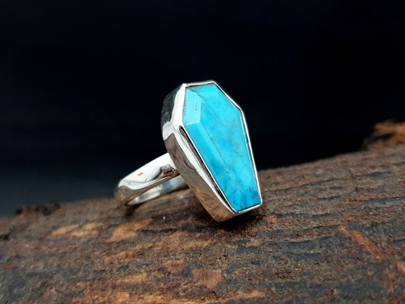K&ouml;nnte beinhalten: Ein silberner Ring mit einem Sarg-f&ouml;rmigen T&uuml;rkisstein. Der hellblaue Stein ist in einem polierten Silberband gefasst und schafft ein einzigartiges Schmuckst&uuml;ck. Der Ring wird auf einer Holzoberfl&auml;che pr&auml;sentiert.