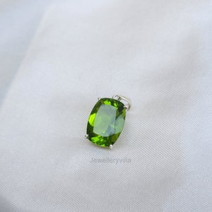 May include: A green peridot gemstone pendant set in silver. The stone is a cushion cut and has a smooth, polished finish.