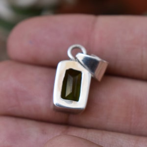 Puede incluir: Un colgante de plata con una piedra preciosa verde rectangular engastada en un engaste de bisel. El colgante tiene un bucle en la parte superior para sujetar una cadena.