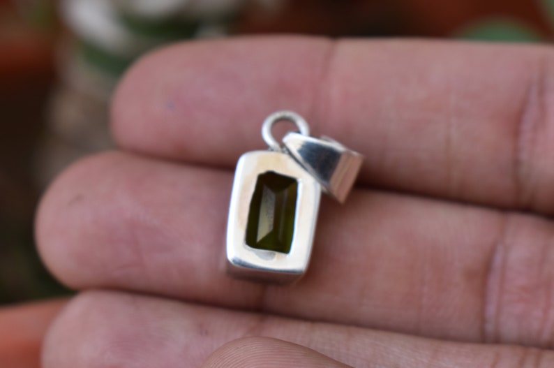 Puede incluir: Un colgante de plata con una piedra preciosa verde rectangular engastada en un engaste de bisel. El colgante tiene un bucle en la parte superior para sujetar una cadena.