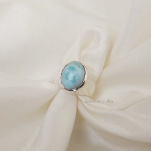 K&ouml;nnte beinhalten: Ein silberner Ring mit einem ovalen blauen und wei&szlig;en Edelstein. Der Ring liegt auf einem wei&szlig;en Stoffhintergrund.