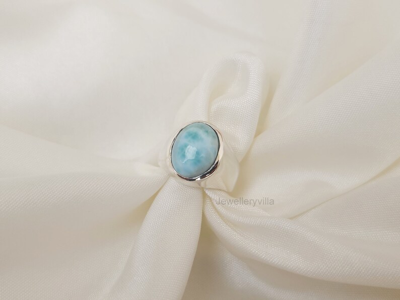K&ouml;nnte beinhalten: Ein silberner Ring mit einem ovalen blauen und wei&szlig;en Edelstein. Der Ring liegt auf einem wei&szlig;en Stoffhintergrund.