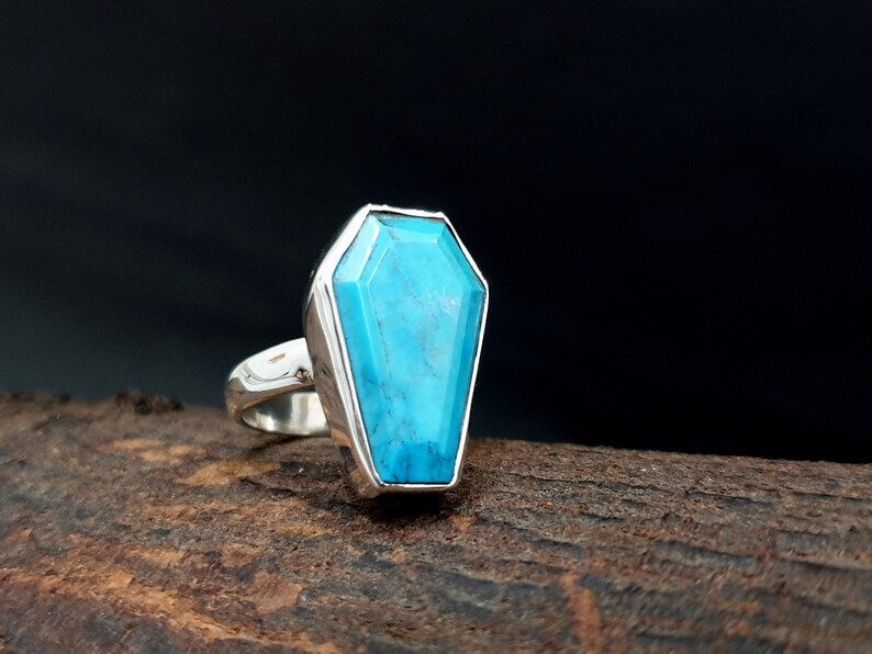 K&ouml;nnte beinhalten: Ein silberner Ring mit einem Sarg-f&ouml;rmigen T&uuml;rkisstein. Der leuchtend blaue Stein ist in eine silberne L&uuml;nette gefasst. Der Ring wird auf einer rustikalen Holzoberfl&auml;che pr&auml;sentiert, was sein einzigartiges Design hervorhebt.