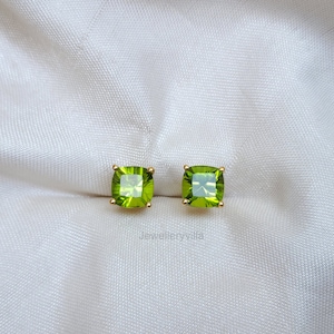 May include: A pair of gold earrings with square green gemstones. The earrings are on a white fabric background.
