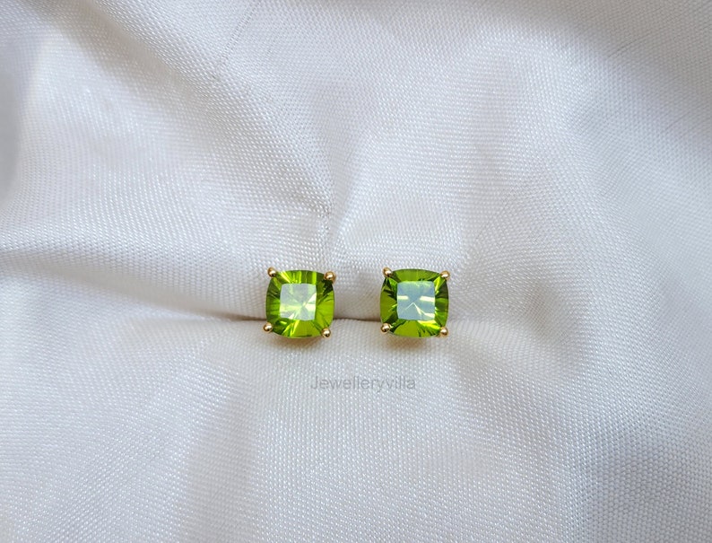 May include: A pair of gold earrings with square green gemstones. The earrings are on a white fabric background.