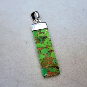 Pu&ograve; includere: Ciondolo rettangolare con una pietra verde e color rame. La pietra &egrave; incastonata in un metallo color argento con un anello nella parte superiore per una catena. Il ciondolo presenta un motivo naturale unico.