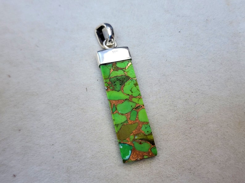 Pu&ograve; includere: Ciondolo rettangolare con una pietra verde e color rame. La pietra &egrave; incastonata in un metallo color argento con un anello nella parte superiore per una catena. Il ciondolo presenta un motivo naturale unico.