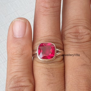 May include: A silver ring featuring a large, square-shaped, bright red gemstone. The ring is worn on a finger.