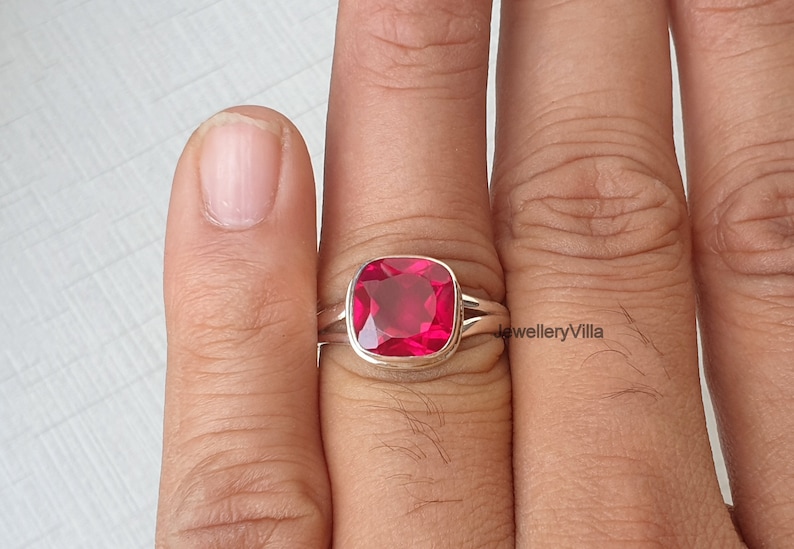 May include: A silver ring featuring a large, square-shaped, bright red gemstone. The ring is worn on a finger.