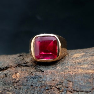 Pu&ograve; includere: Un anello in oro con una grande pietra preziosa di colore rosso vibrante a taglio quadrato, incastonata in una fascia di metallo liscia e lucida. L'anello &egrave; posizionato per evidenziare il colore cremisi profondo e la superficie sfaccettata.