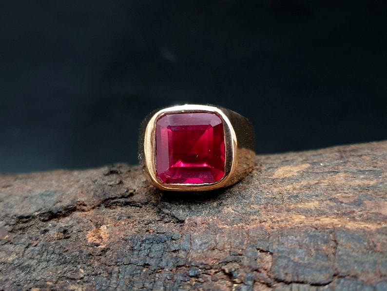 Pu&ograve; includere: Un anello in oro con una grande pietra preziosa di colore rosso vibrante a taglio quadrato, incastonata in una fascia di metallo liscia e lucida. L'anello &egrave; posizionato per evidenziare il colore cremisi profondo e la superficie sfaccettata.