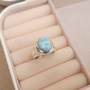 K&ouml;nnte beinhalten: Ein silberner Ring mit einem hellblauen ovalen Edelstein. Der Ring befindet sich in einer wei&szlig;en Ringbox mit beigefarbenem Samtbezug.