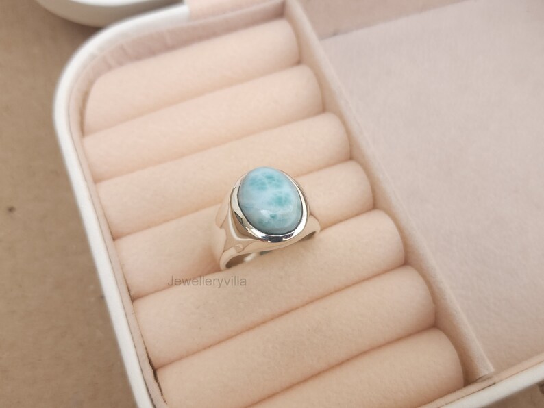 K&ouml;nnte beinhalten: Ein silberner Ring mit einem hellblauen ovalen Edelstein. Der Ring befindet sich in einer wei&szlig;en Ringbox mit beigefarbenem Samtbezug.