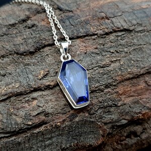 May include: A silver chain necklace with a pendant shaped like a coffin. The pendant features a large, faceted, blue gemstone.