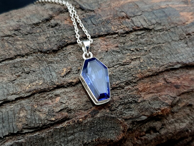 May include: A silver chain necklace with a pendant shaped like a coffin. The pendant features a large, faceted, blue gemstone.