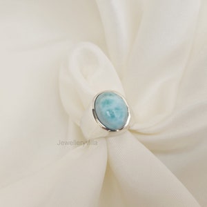 K&ouml;nnte beinhalten: Ein silberner Ring mit einem ovalen, hellblauen Edelstein. Der Ring liegt auf einem wei&szlig;en Stoffhintergrund.