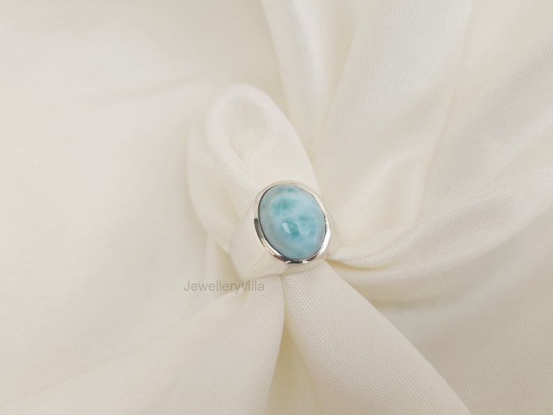 K&ouml;nnte beinhalten: Ein silberner Ring mit einem ovalen, hellblauen Edelstein. Der Ring liegt auf einem wei&szlig;en Stoffhintergrund.