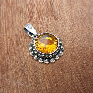May include: A silver pendant featuring a round, faceted, amber-coloured gemstone set in a silver bezel. The bezel is surrounded by a decorative border of small silver beads.