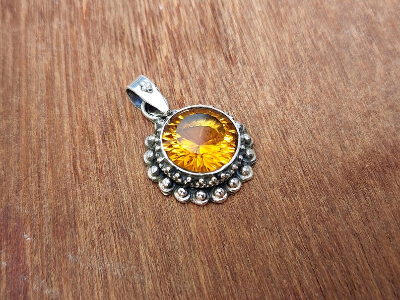 May include: A silver pendant featuring a round, faceted, amber-coloured gemstone set in a silver bezel. The bezel is surrounded by a decorative border of small silver beads.
