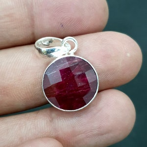 Puede incluir: Un colgante redondo de piedra roja con una argolla de plata. La piedra preciosa es facetada y tiene un color rojo intenso.