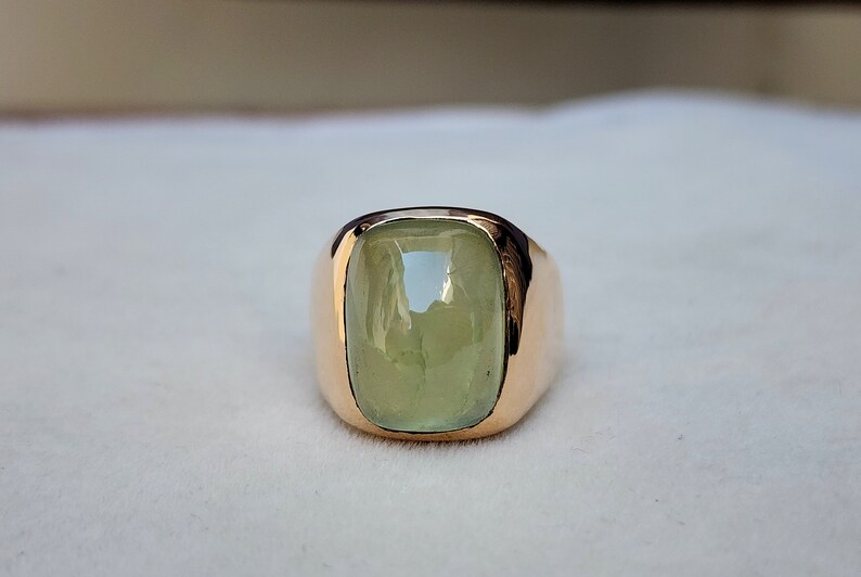 Puede incluir: Un anillo de oro con una gran piedra preciosa verde claro engastada en una forma cuadrada.