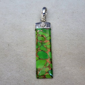 Pu&ograve; includere: Ciondolo rettangolare con anello argentato e pietra verde e ramata vibrante. La pietra presenta un motivo strutturato unico. Il ciondolo &egrave; lungo circa 5 cm, mostrando una miscela di colori e trame naturali.