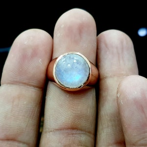 Puede incluir: Un anillo con una gran piedra de luna ovalada engastada en una banda dorada. La piedra tiene un color blanco lechoso con un ligero brillo iridiscente.