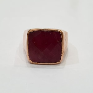 May include: A square-shaped ring with a large, deep red gemstone set in a gold-toned metal band.