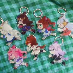以下が含まれることがあります： アニメスタイルのキャラクターキーホルダーのコレクション。各キーホルダーは、鮮やかな色と詳細を備えたユニークなキャラクターデザインが特徴で、銀色のキーリングに取り付けられています。キーホルダーは、緑と白のチェック柄の生地に表示されています。
