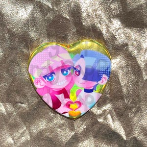 May include: Heart-shaped pin featuring two anime-style characters with pink and blue hair, embracing. The pin has a gold border and a holographic effect. The characters are holding a heart-shaped object.