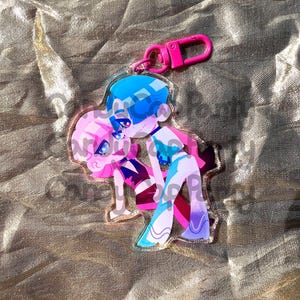 May include: A clear acrylic keychain featuring two cartoon figures in shades of pink and blue, with a pink clasp. The figures are embracing. The keychain has a gold border.