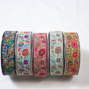Op de afbeelding: Zes spoelen decoratief lint met bloemenborduurwerk in verschillende kleuren. De linten hebben een reeks kleuren, waaronder marineblauw, roze en groen, met ingewikkelde bloempatronen. De spoelen zijn naast elkaar geplaatst.