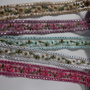 Könnte beinhalten: Fünf Streifen dekoratives Stoffband mit Blumenstickerei und kleinen runden Spiegeln. Die Bänder zeigen verschiedene Farben wie Gold, Mauve, Hellblau, Weiß und leuchtendes Pink, jeweils mit grünen Blattakzenten und gewellten Rändern.