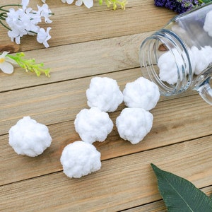 Op de afbeelding: Een close-up van zes witte, pluche ballen op een houten oppervlak. De ballen zijn in een halve cirkel gerangschikt, met &eacute;&eacute;n bal die lichtjes overlapt met een andere. De ballen zijn zacht en lijken gemaakt te zijn van katoen of wol.