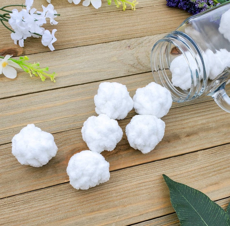 Op de afbeelding: Een close-up van zes witte, pluche ballen op een houten oppervlak. De ballen zijn in een halve cirkel gerangschikt, met &eacute;&eacute;n bal die lichtjes overlapt met een andere. De ballen zijn zacht en lijken gemaakt te zijn van katoen of wol.