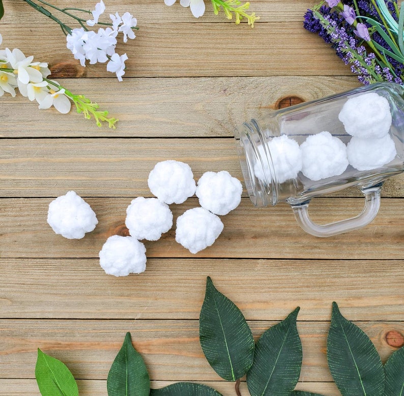 Op de afbeelding: Witte wattenbolletjes die uit een glazen pot op een houten oppervlak met groene bladeren en witte bloemen vallen.